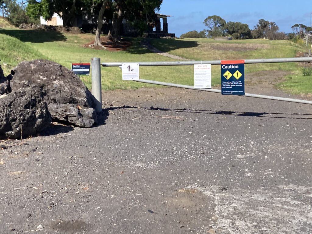 A picture of a gravel driveway with a metal gate. The gate contains four signs. There is a volcanic rock or lava bomb on the left of the picture and trees in the distance.