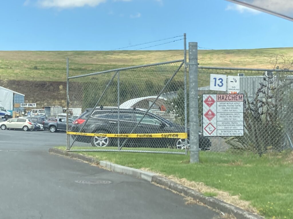 A photo looking up a driveway of a factory with a small volcano in the background. There is a fence and cars in the foreground.